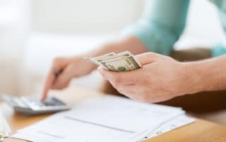 A closeup of a person sitting at a desk with financial documents in front of them, typing on a calculator with one hand while holding money in the other.